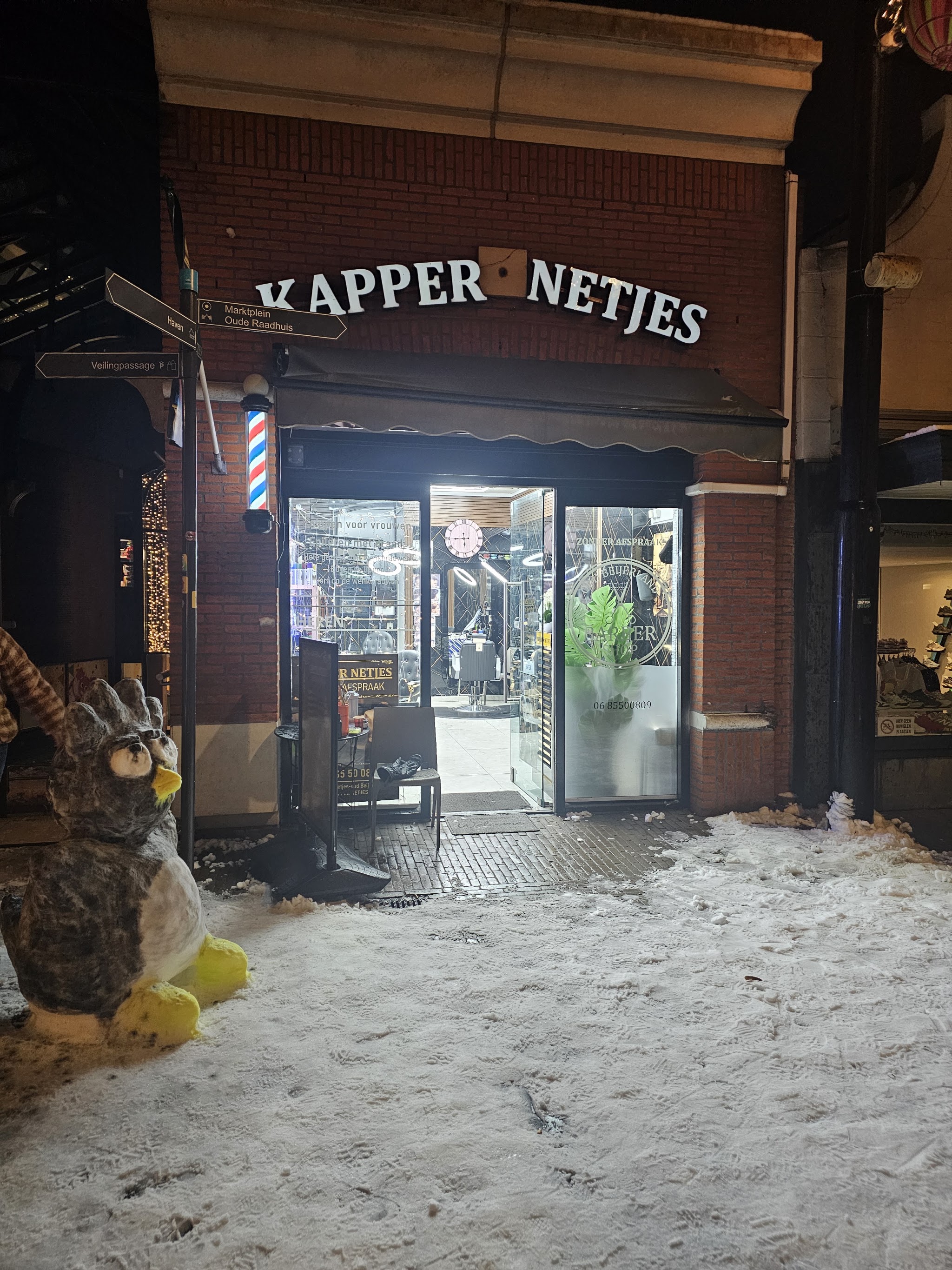 Kapper Netjes bij avond: de verlichte etalage, het bord KAPPER NETJES en de barbierspaal, met sneeuw op de stoep
