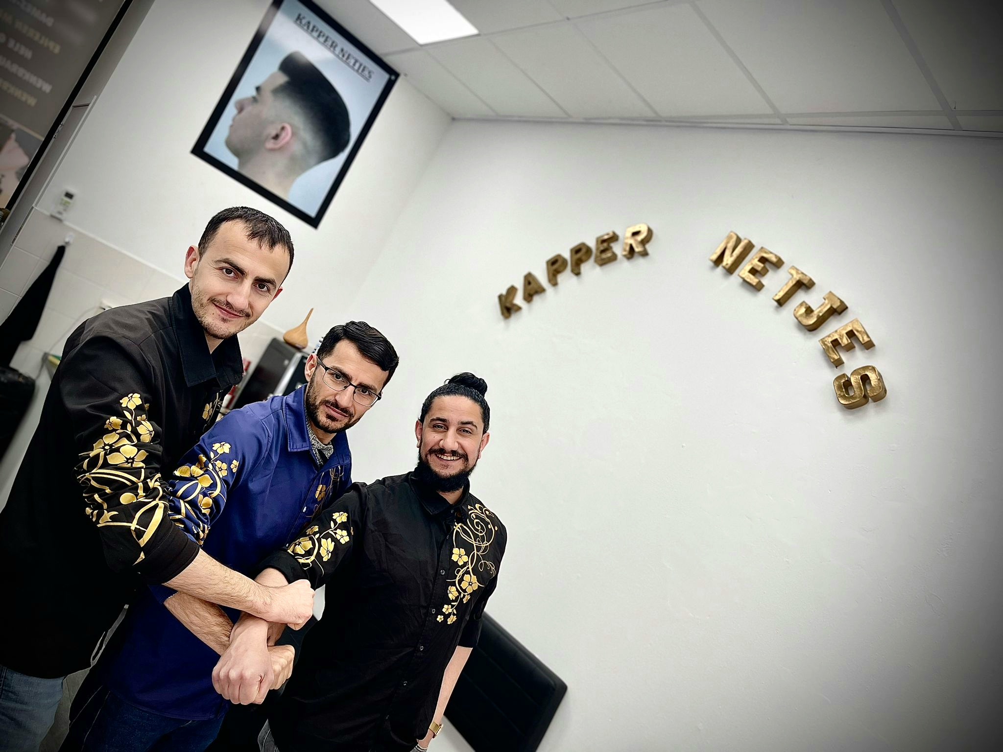 Drie kappers van Kapper Netjes samen onder de gouden letters KAPPER NETJES op de muur